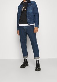 Giacca di jeans blu scuro con colletto in shearling, felpa nera sotto, jeans blu arrotolati e stivaletti neri con suola spessa.