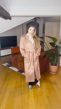 Ženska z dolgimi lasmi, oblečena v roza trenč plašč, šal z vzorcem, črne hlače in srebrne čevlje, stoji v dnevni sobi z rastlino in televizorjem.