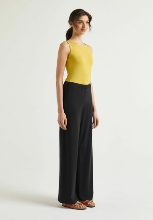 Donna in piedi con top giallo senza maniche, pantaloni neri a gamba larga e sandali marroni su uno sfondo chiaro uniforme.