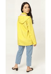 Chaqueta impermeable amarilla con capucha, con un forro interior de diseño, combinada con jeans de color azul claro y sandalias negras de tacón cuadrado.