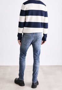 Marinblå och vit randig tröja med långa ärmar, kombinerad med ljusblå jeans och svarta skor. Har en rund halsringning och en figurnära design.