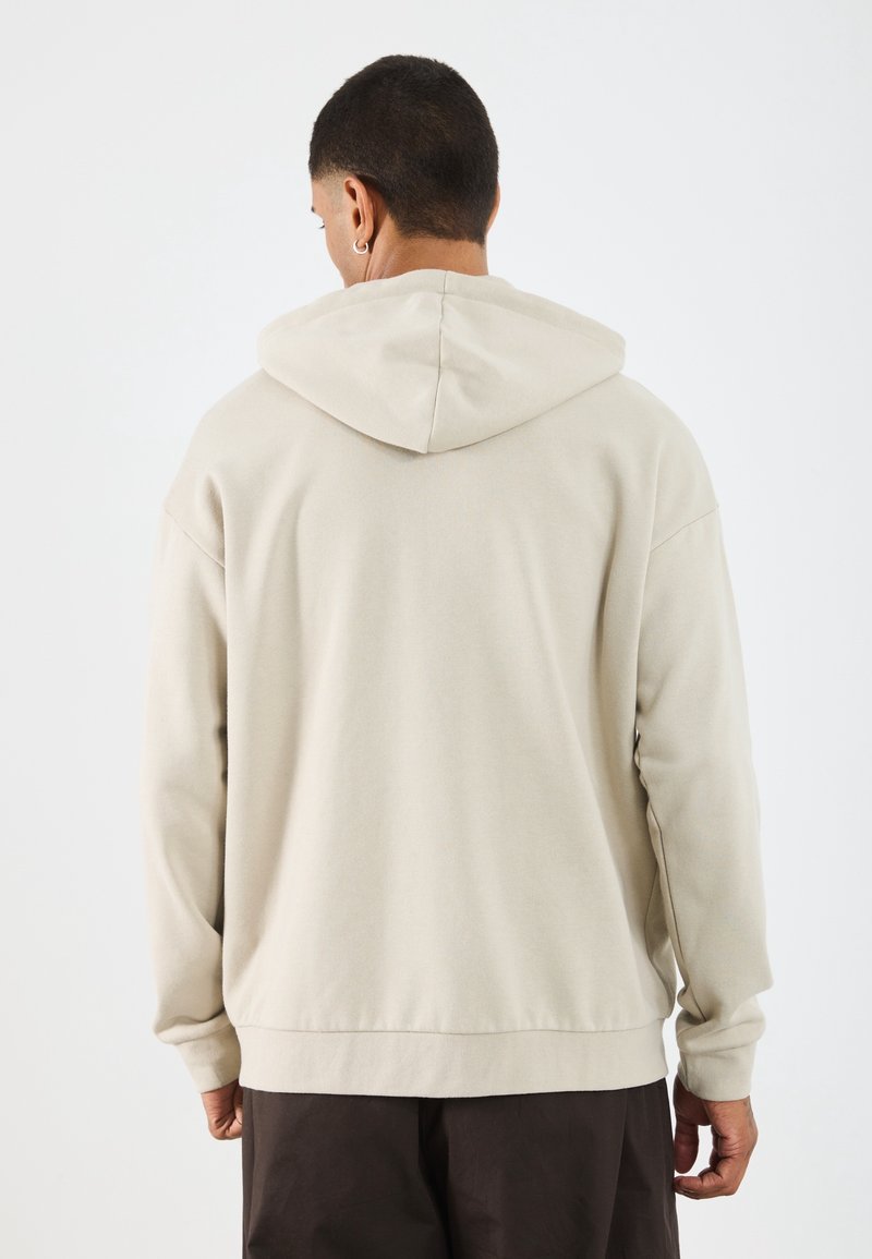 Homme portant un sweat à capuche beige et un pantalon sombre, vu de dos sur un fond clair uni.