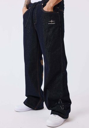 Personne portant un jean large bleu foncé avec le logo PXP et des baskets blanches, les mains dans les poches, debout devant un fond uni.
