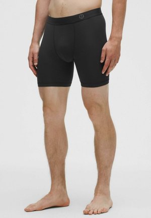 Schwarze Boxershorts aus Stoff mit glatter Textur, mittellang bis zum Oberschenkel, und einem Bund mit dezentem Logo. Keine Muster.