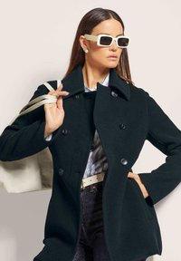 Black wool coat with a double-breasted design, large collar, and black buttons; paired with a light blue striped shirt and a beige handbag.