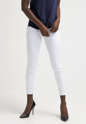 Persoon met witte skinny jeans, een marineblauw mouwloos topje en zwarte hoge hakken, staand tegen een effen achtergrond.
