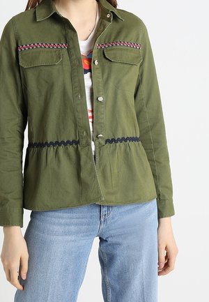 Femme portant une veste ouverte vert olive avec des bandes décoratives, un t-shirt blanc graphique et un jean bleu clair sur fond blanc.