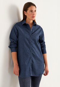 Tummansinisestä denimhousupaidasta, jossa on nappikiinnitys, pitkät hihat, jotka on käännetty hihoista, ja kaareva helma. Tasainen rakenne, klassinen kaulustyyli.