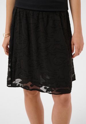 Jupe noire longueur genou avec motifs floraux texturés portée par une personne aux jambes nues et bracelets dorés au poignet gauche.