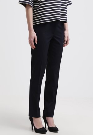 Donna che indossa pantaloni neri aderenti, tacchi alti neri e una maglietta a maniche corte a righe bianche e nere, in piedi davanti a uno sfondo grigio uniforme.