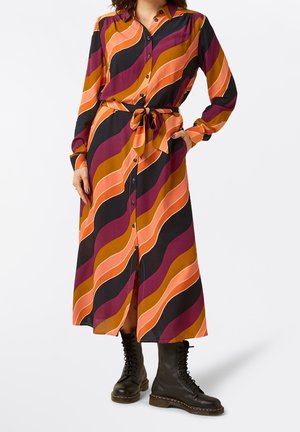 Woman wearing a long-sleeve, buttoned, belted midi dress with wavy stripes in orange, black, purple, and brown, paired with black lace-up boots.