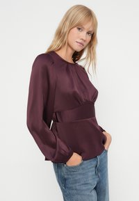 Satiini, viininpunainen pusero, jossa on gathered-kaula-aukko ja istuva vyötärö. Pitkät hihat elastisilla cuffeilla, yhdistettynä sinisiin farkkuihin.