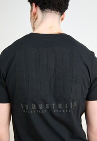 Musta puuvillapaita, jossa on suuret kohokuvioidut tekstit "ALPHA" ja "INDUSTRIES" kontrastiväreissä. Pyöreä kaula-aukko ja lyhyet hihat.