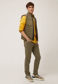 Chaleco acolchado ligero de color verde oliva sobre una camisa de manga larga amarilla, combinado con pantalones ajustados de color oliva y zapatillas beige con acentos azules.