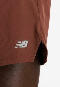 Pantalones cortos deportivos marrones con textura perforada. Presentan un logotipo blanco de New Balance cerca del dobladillo y aberturas laterales para facilitar el movimiento.