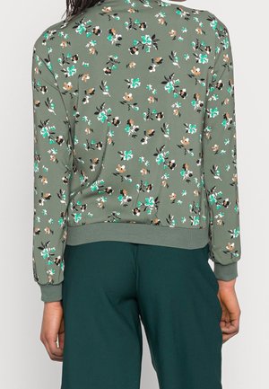 Back view of person wearing green floral long-sleeve top with ribbed cuffs and dark green pants.