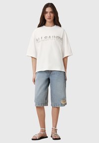 Camicetta bianca a maniche corte con la scritta "ALLSAINTS" in lettere testurizzate. Abbinata a pantaloni corti in denim azzurri con orlo strappato e sandali.