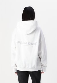 Sweat à capuche blanc avec une grande impression grise de "PEGADOR" dans le dos. Coupe ample avec capuche à cordon et poignets côtelés. Texture de tissu douce.