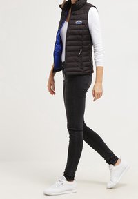 Svart quiltad väst med blå insida, dragkedjefickor och en logotypapplikation. Parat med smala svarta jeans och vita sneakers.