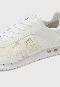White athletic shoe with mesh and faux leather materials, featuring gold accents, textured sole, and white laces. Logo appears on side and tongue.
