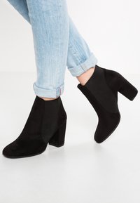 Stivali alla caviglia neri in morbido suede, con tacco spesso e pannelli laterali elastici, abbinati a jeans azzurri arrotolati.