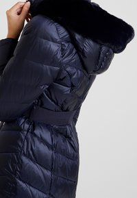Chaqueta tipo puffers de color navy con un diseño acolchado, cuello de piel sintética, cintura ceñida con cinturón y textura suave y brillante. Presenta una silueta ajustada.