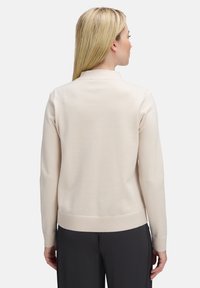 Vrouw met lang blond haar, gekleed in een lichtbeige trui en zwarte broek, met haar rug naar de camera gekeerd tegen een effen witte achtergrond.