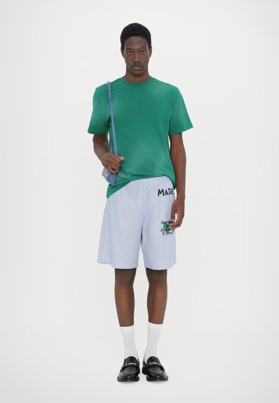 T-shirt verde in cotone, pantaloni corti blu a righe con testo e grafiche ricamati, abbinati a scarpe nere e calzini bianchi. Borsa a spalla nera.