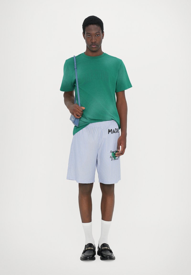 T-shirt verde in cotone, pantaloni corti blu a righe con testo e grafiche ricamati, abbinati a scarpe nere e calzini bianchi. Borsa a spalla nera.