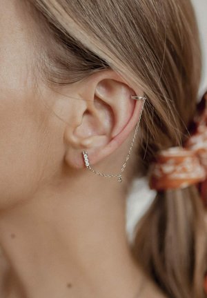 Silberner Ohrcuff und Kette mit kleinen klaren Edelsteinen, die mit einer filigranen Kette am Ohrläppchen befestigt sind. Glatte Metalloberfläche, minimalistisches Design.