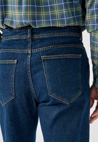 Jeans in denim blu scuro con cuciture a vista, due tasche posteriori con pattine a punta e un taglio a vita media. Indossati con una camicia a quadri.