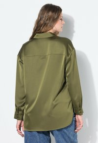 Donna con capelli lunghi che indossa una camicia di satin a maniche lunghe verde oliva e jeans blu, di spalle contro un muro bianco.