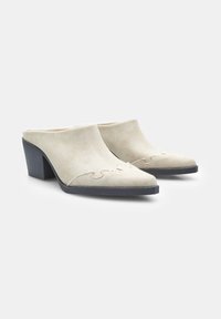 Mule kain beige dengan permukaan bertekstur, ujung runcing, dan pola jahitan bergelombang; didukung oleh tumit blok hitam yang kontras.