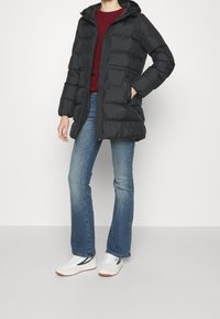 Černá prošívaná bunda s kapucí, s zipovým uzávěrem, horizontálním prošíváním a předními kapsami, nošena přes červený svetr a džíny.