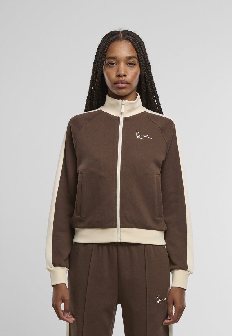 Brown zip-up jacket with cream accents, featuring a high collar, front pockets, and ribbed cuffs. The fabric appears soft and textured.