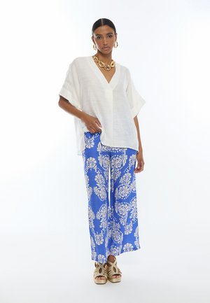 Mujer de pie lleva un top blanco suelto de cuello en V, pantalones anchos azules con estampado floral blanco, joyas doradas y sandalias de plataforma tejidas.
