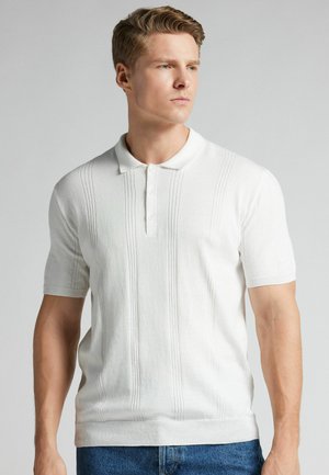 Mann mit kurzen blonden Haaren, der ein weißes Poloshirt mit kurzen Ärmeln und blaue Jeans trägt, leicht nach rechts gewandt vor einem schlichten Hintergrund.