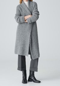 Persona vestida con un largo cárdigan gris texturizado sobre pantalones gris con puños y botas de cuero negras hasta el tobillo, de pie con los brazos cruzados.