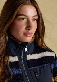Fleece jacket with dark blue, cream, and tan stripes; high collar; zip closure; soft texture; features zippered pockets.