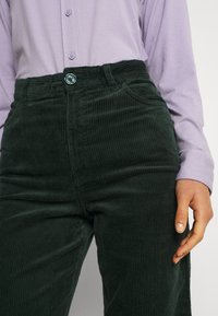 Gröna flätade byxor med en strukturerad yta, hög midja och fickor framtill. Kombination med en ljuslila långärmad skjorta.