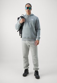 Grå hettegenser med lommer foran og logo, kombinert med grå joggebukser. Svart ryggsekk og overdimensjonerte reflekterende solbriller fullfører stilen.