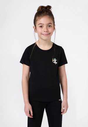 Fille aux cheveux bruns en chignon, portant un t-shirt noir avec un petit logo coloré et un pantalon noir assorti, se tenant contre un fond blanc.