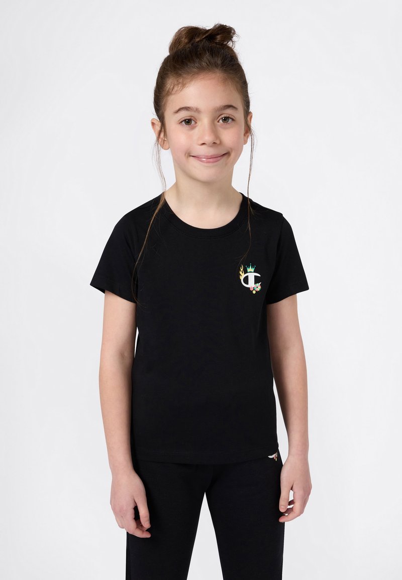 Κορίτσι με καστανά μαλλιά σε αλογοουρά, φορώντας μαύρη μπλούζα με ένα μικρό πολύχρωμο λογότυπο και αντίστοιχες μαύρες παντελόνες, στέκεται μπροστά σε λευκό φόντο.