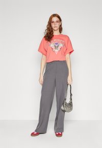 Coral graphic t-shirt featuring eagle design, gray high-waisted wide-leg trousers, and red shoes with gray accents, holding a metallic handbag.