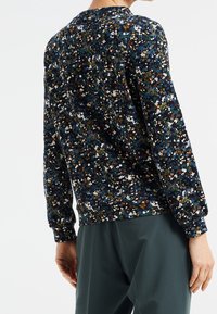 Donna che indossa una blusa a maniche lunghe a fiori scuri con polsini arricciati e pantaloni verde scuro, mostrata da dietro.