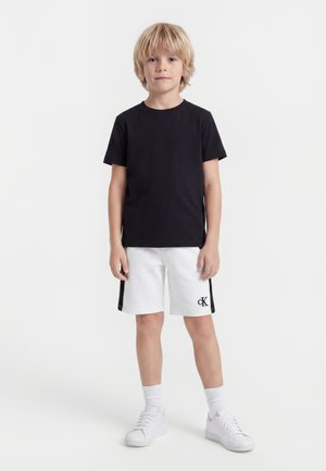 Noor poiss seisab näoga ettepoole, seljas must t-särk, valged lühikesed püksid külgtriipudega ja valged tossud, taust valge.