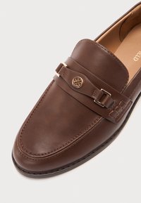 Mocasín de cuero marrón con textura suave, punta redondeada, detalle de costura y una correa decorativa con un acento dorado.