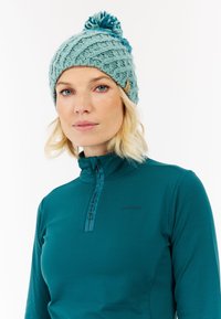 Světle tyrkysová pletená čepice s texturovaným vzorem, s bambulí na vrchu a malou cedulkou. Nosená s odpovídajícím tyrkysovým zipovým svetrem.