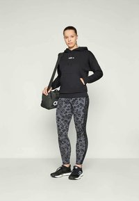 Sweatshirt preto com capuz e um pequeno logótipo, combinado com leggings cinzas em padrão de leopardo com riscas laterais. Os acessórios incluem uma mala de ginásio preta.