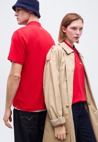 Άνδρας με κόκκινο πόλο και ναυτικό καπέλο τύπου κάπα στέκεται πλάτη με πλάτη με γυναίκα που φορά μπεζ παλτό και κόκκινο τοπ, και οι δύο με σκούρο τζιν, ουδέτερο φόντο.